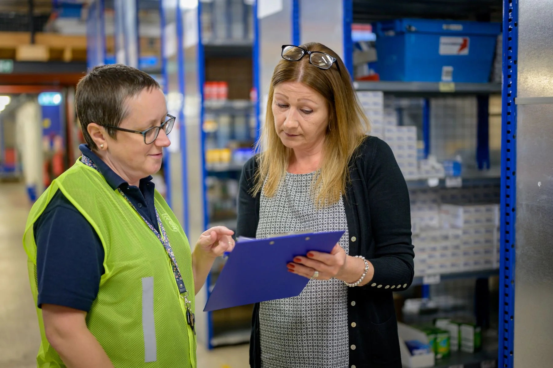 Two people discussing a clipboard near warehouse shelving